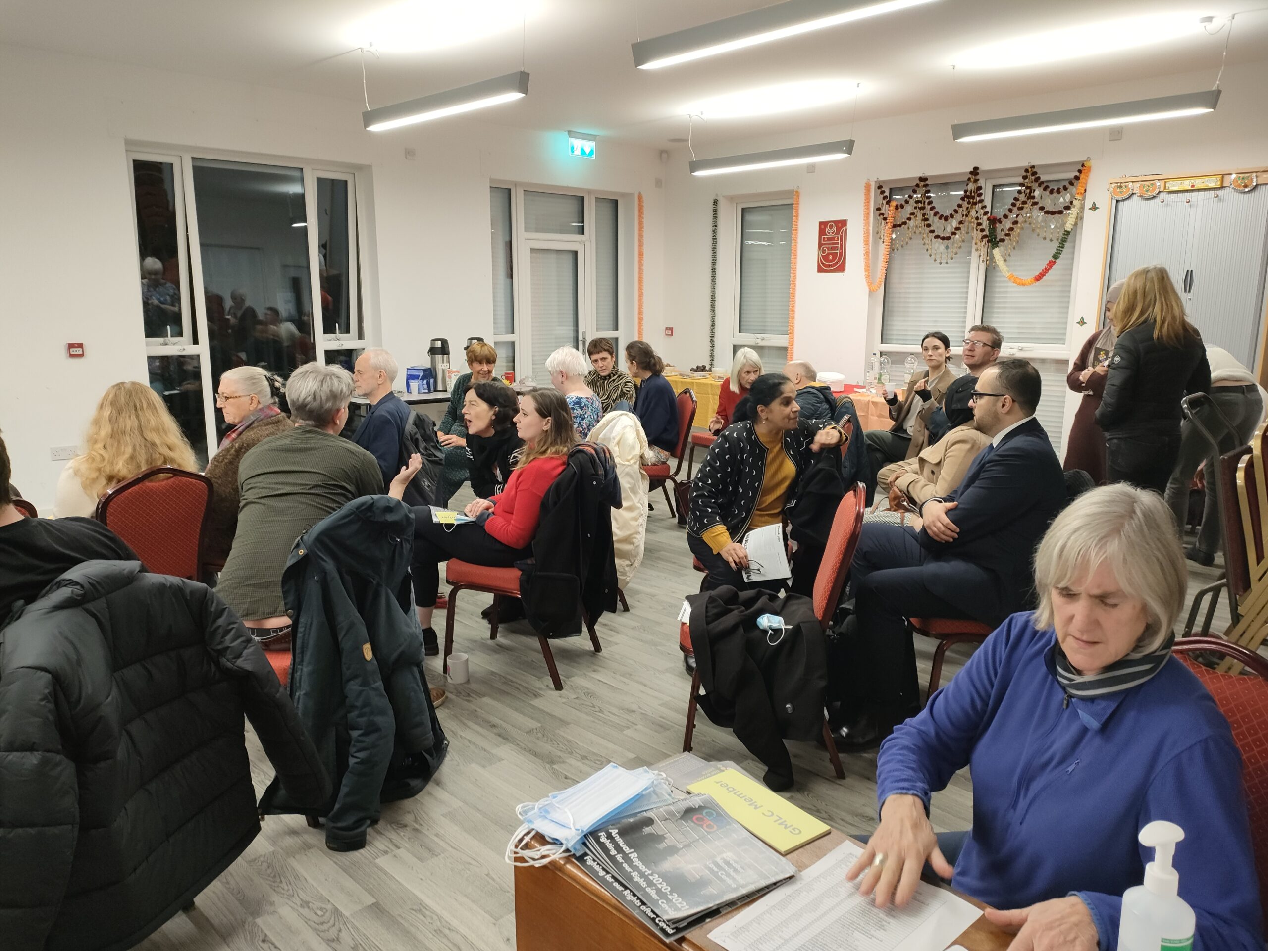 Image shows a full room at a GMLC AGM, with a volunteer in the foreground signing people in and giving out agendas and reports.