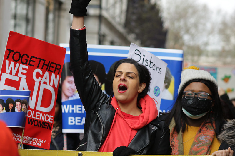 Image shows a woman of colour, arm raised, at a protest march, next to a placard that says 'Sisters' and another that says 'together we can end violence against women'.