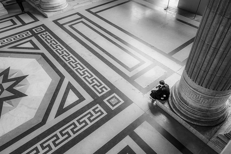 B&W image shows a lone person sitting against a column in a large, grand court hallway.