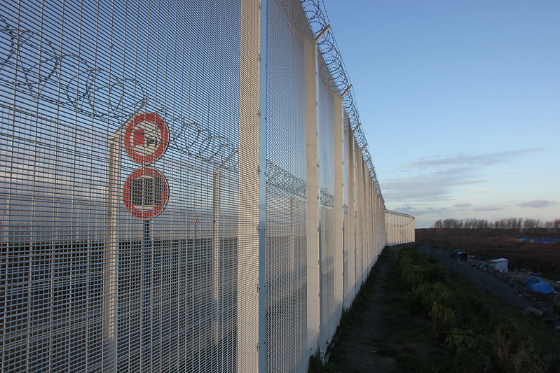 Image shows a fence at least 10 foot high, with scrub land in the foreground on the right, and behind the fence, a series of warning signs.