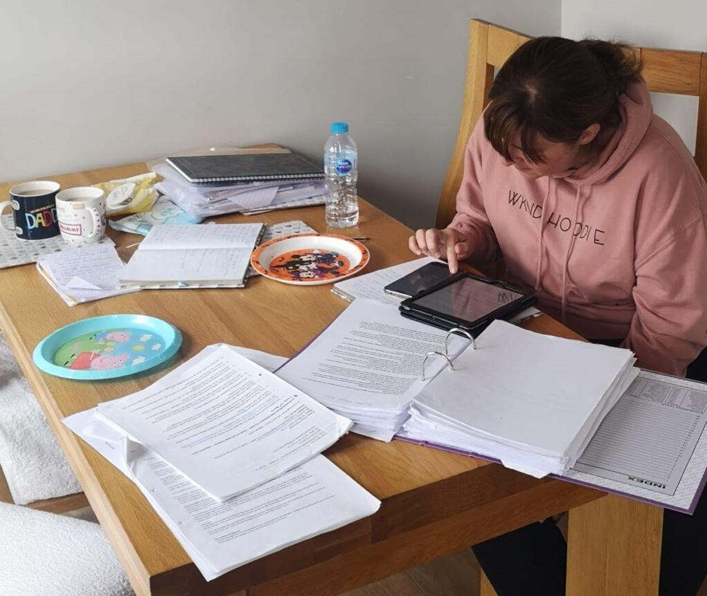 Lisa Murray, head down and wearing a pink sweatshirt, leans over a table covered in paperwork, empty plates, cups and a water bottle while preparing her bundle for a Tribunal hearing.
